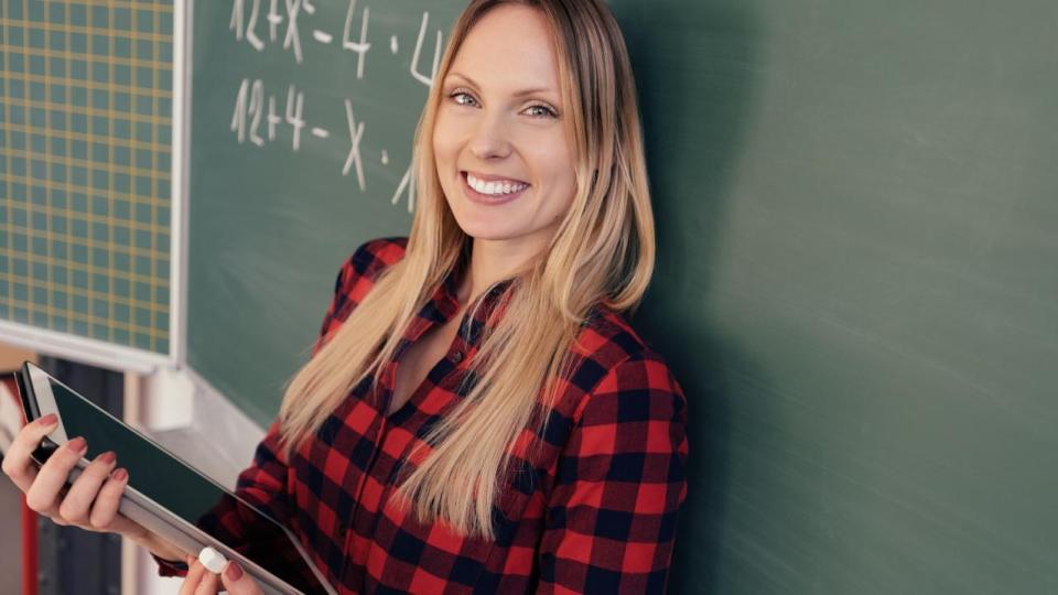 Eine Lehrerin steht mit dem Rücken zur Tafel, in ihren Händen hält sie ein Tablet-Computer.