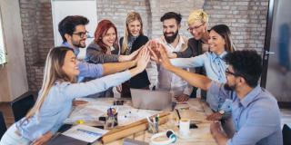 Gruppe Erwachsener lehnt sich über einen Arbeitstisch und bringt die Hände in der Mitte zusammen.