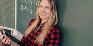 Eine Lehrerin steht mit dem Rücken zur Tafel, in ihren Händen hält sie ein Tablet-Computer.